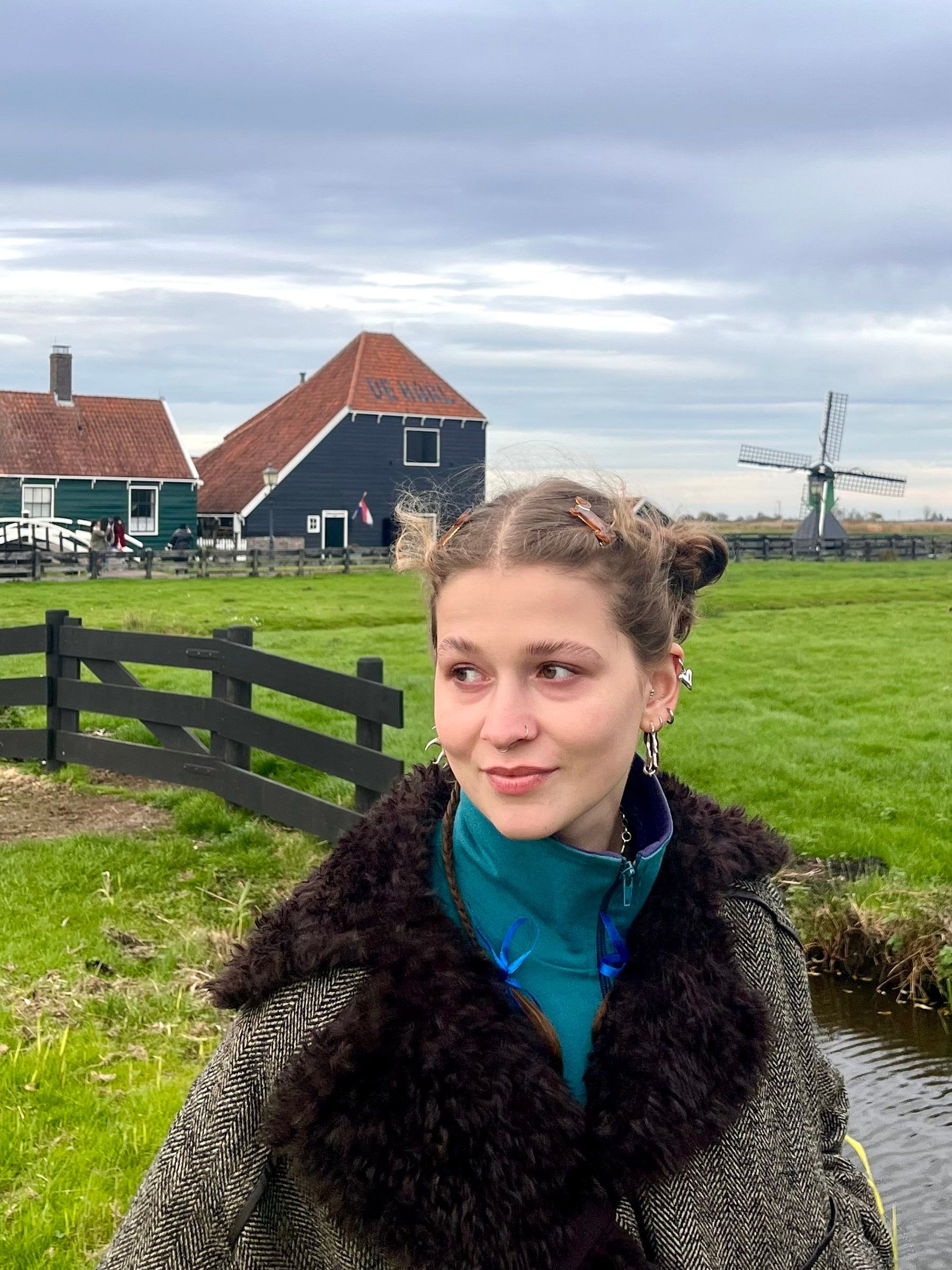 Mila Wójcicka outdoors, looking to the side, with wooden houses and a windmill in the background.