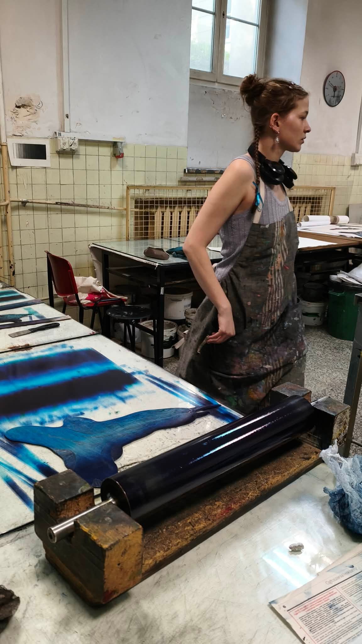 Mila in a printmaking workshop, standing behind a table with an ink roller and a large print in blue ink.